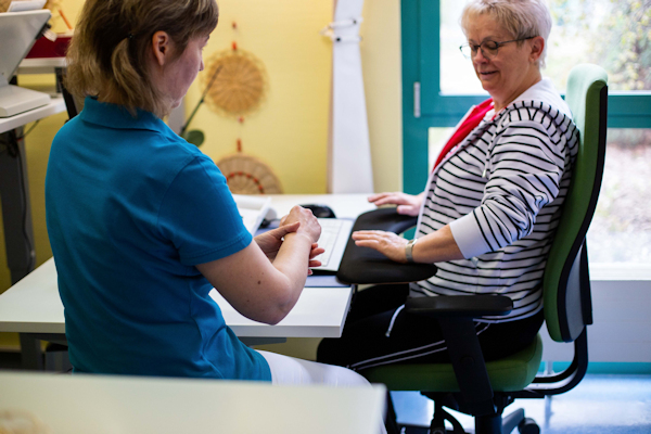 Therapeutin erklärt einer am Schreibtisch sitzenden Rehabilitandin die ergonomische Handhaltung  Therapeutin erklärt einer am Schreibtisch sitzenden Rehabilitandin die ergonomische Handhaltung
